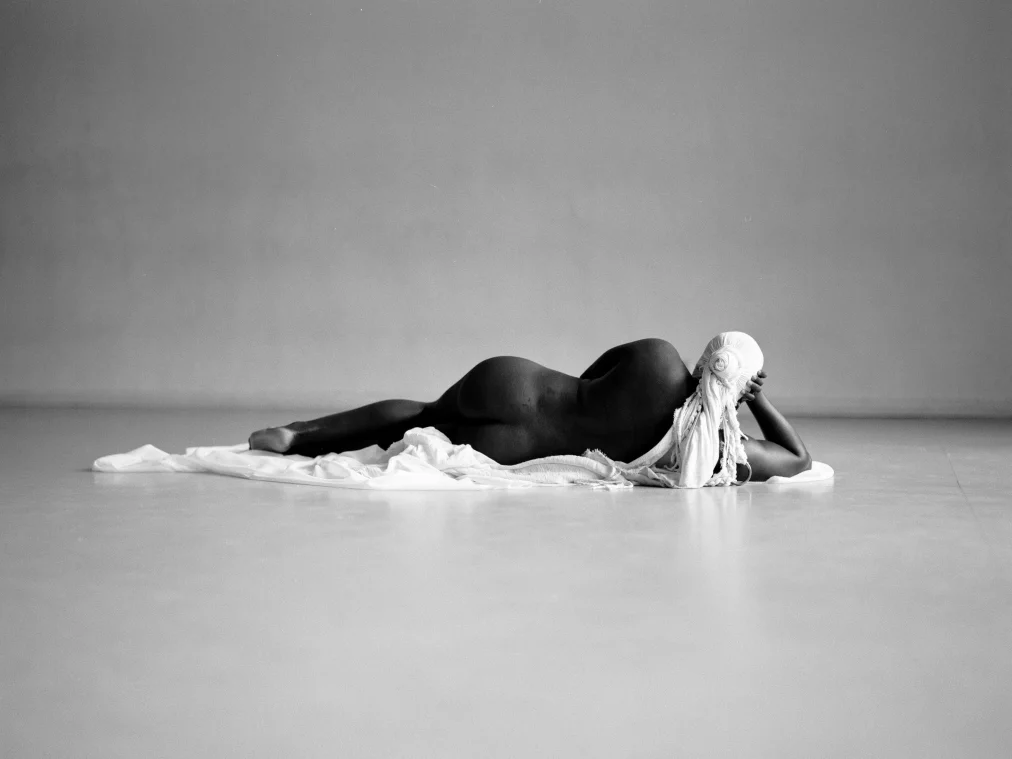 A person lies naked on their stomach on a white cloth in an empty room. Their head is covered with a white fabric.