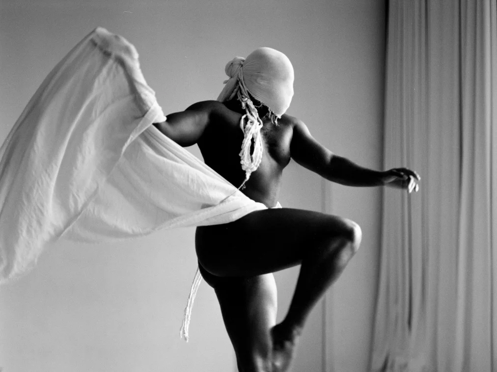 A standing person, naked, lifts one leg and holds a large white cloth. Their head is wrapped in white fabric.