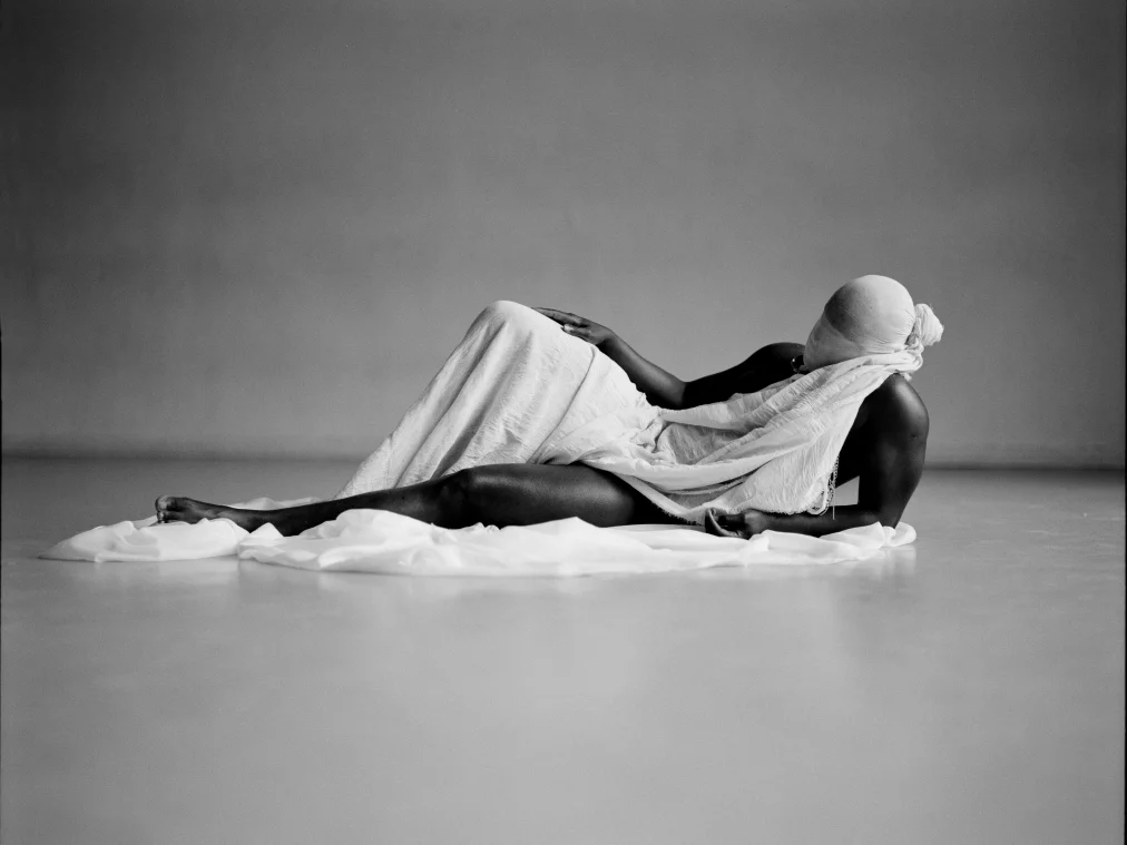 A person lies sideways on a white cloth in an empty room. Their body is partially covered with a white fabric, and their head is fully wrapped in cloth.