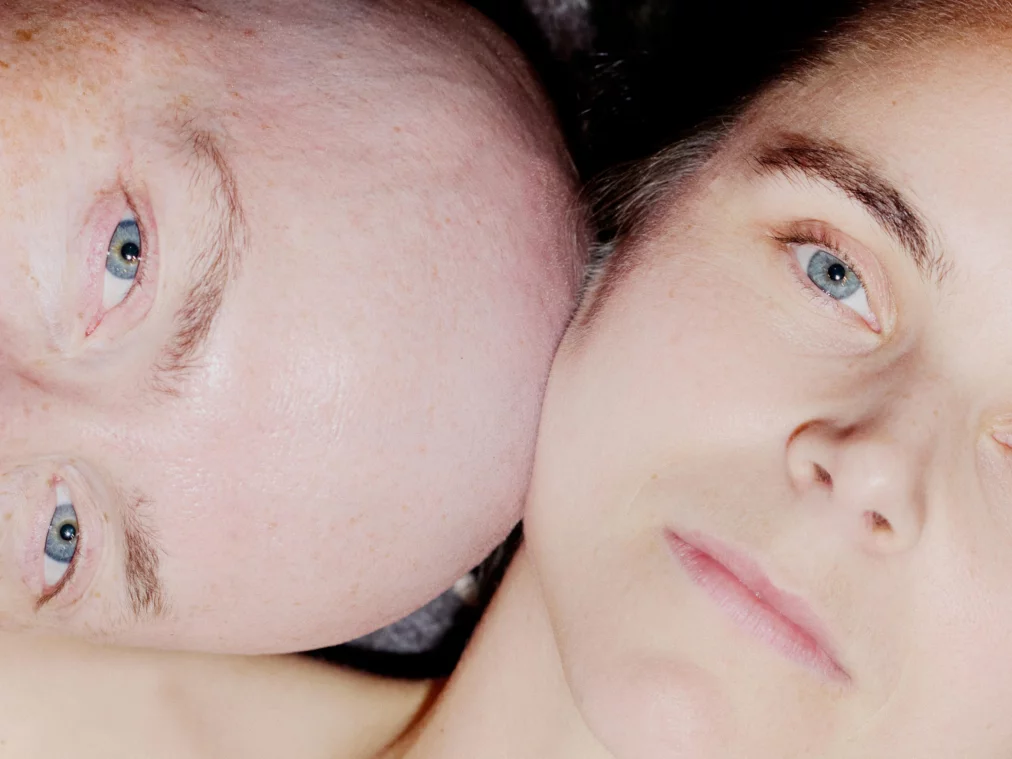 Close-up of two faces lying closely together. One head rests sideways, the other faces forward. Both look ahead with open eyes, their skin highlighted by light reflections.