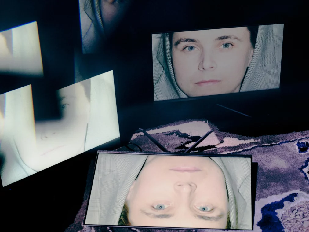 Drei Bildschirme auf einem gemusterten Teppich zeigen in unterschiedlichen Positionen dasselbe Gesicht mit Kapuze und blauen Augen. Der Raum ist dunkel, die Gesichter wirken überbelichtet.