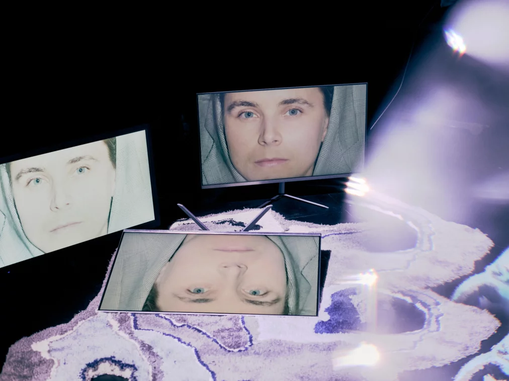 Drei Monitore auf einem Teppich zeigen frontal das Gesicht einer Person mit Kapuze und blauen Augen. Im Vordergrund verwischen Lichtreflexe die Sicht.