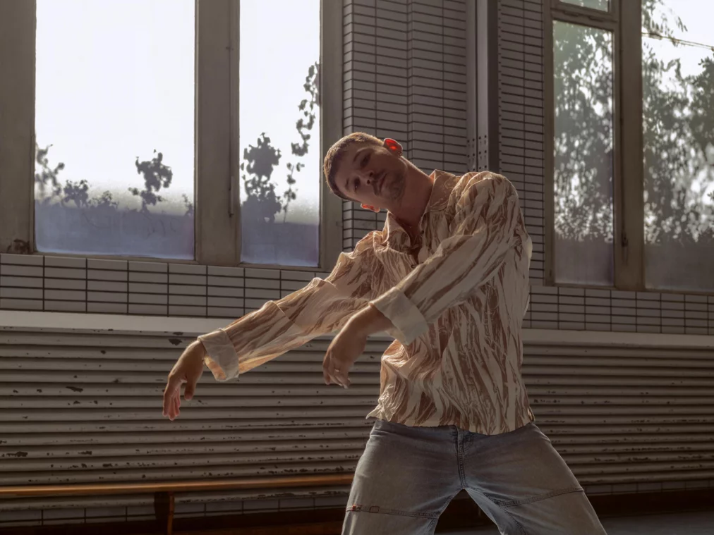 A person wearing a light patterned blouse and jeans poses in front of large windows with plant shadows visible. Their arms are lifted in a curved position, the head tilted to the side, with a calm gaze directed forward.