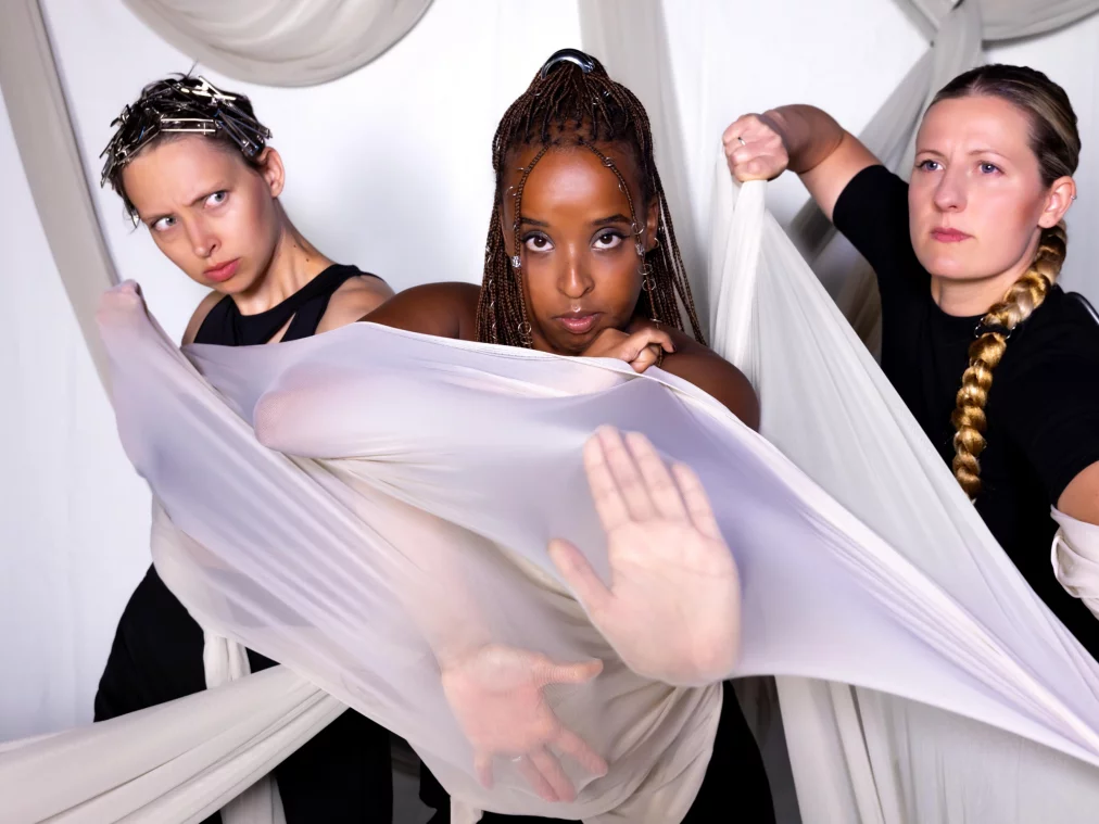 Three performers pose powerfully against a white backdrop. On the left, a person with short hair and many metallic hair clips gazes forward with determination. In the center, a person with long braids looks directly into the camera with a serious expression, extending their hand in a defensive gesture. On the right, a person with a stern expression and a long braided ponytail holds a piece of fabric firmly. All three are entangled in sheer, light fabric that wraps around their bodies.