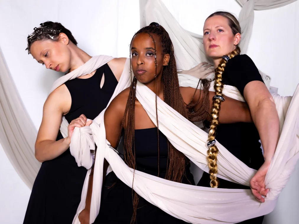Three performers are wrapped in light fabric strips connecting their bodies. On the left, a person with clips in short hair tilts their head downward. In the center, a person with long braids looks forward, fabric draped diagonally across their torso. On the right, a person with a long braid gazes determinedly into the distance, pulling the fabric firmly.