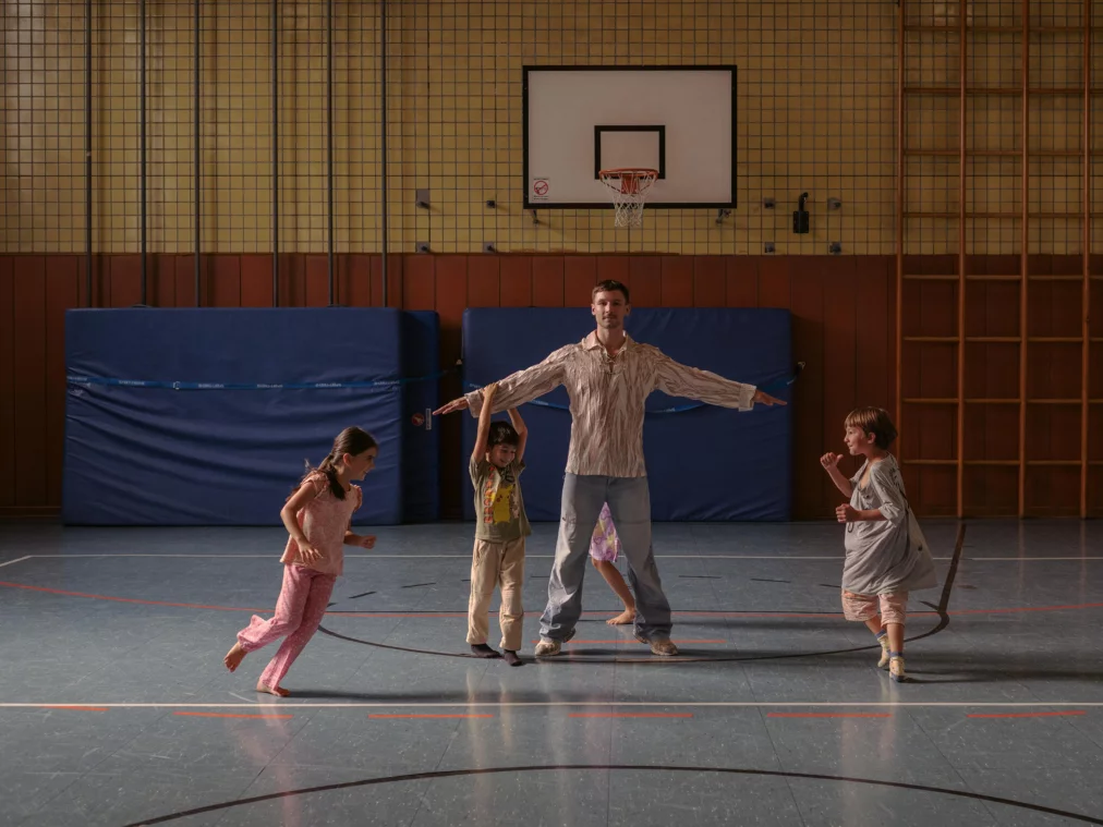 Eine Person steht mit ausgebreiteten Armen in der Mitte einer Turnhalle, während mehrere Kinder um sie herum spielen und laufen. Die Szene ist eingebettet in eine alltägliche Sporthallenumgebung mit blauen Matten, einer Sprossenwand und einem Basketballkorb im Hintergrund.. 
