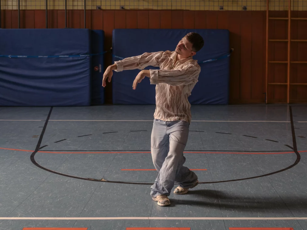 Eine Person tanzt allein in einer Turnhalle. Sie trägt eine helle, gemusterte Bluse und weite Jeans. Die Arme sind nach vorne und seitlich abgewinkelt, der Kopf ist leicht geneigt, als würde die Person in eine fließende Bewegung übergehen.