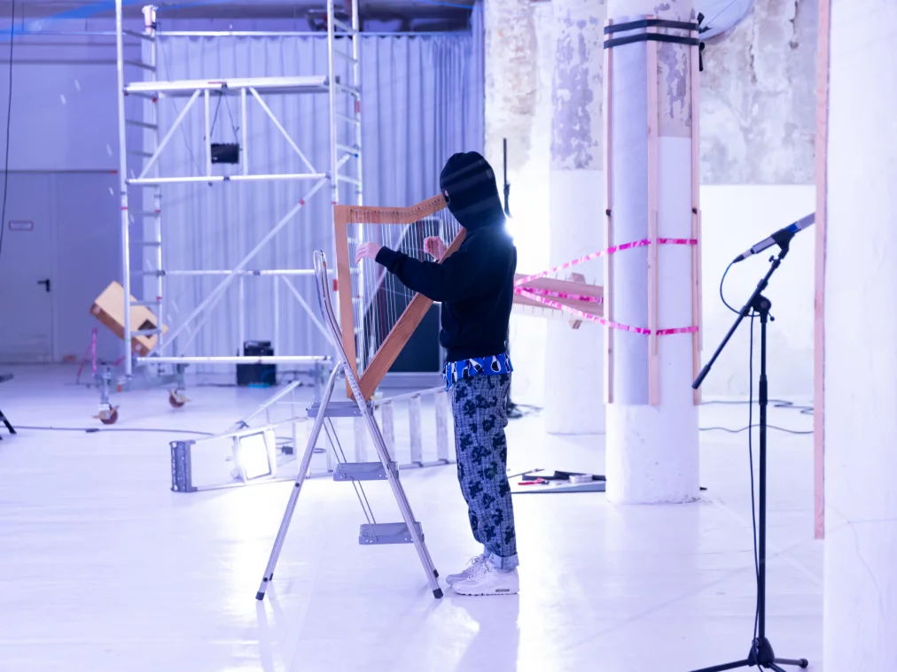 ine Person steht auf einer Leiter und spielt an einer Harfe aus Holz. Sie trägt einen schwarzen Kapuzenpullover und gemusterte Hose. Im Hintergrund sind Säulen, Gerüste und technische Geräte in einem hellen Raum zu sehen.