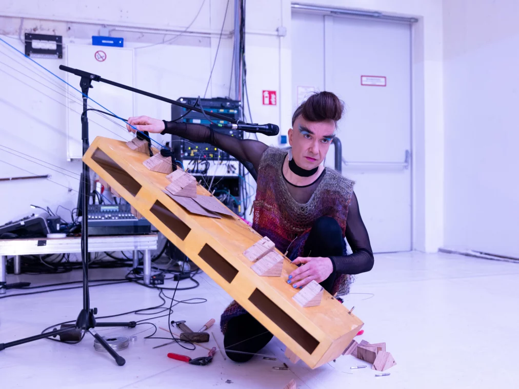 Eine Person mit auffälligem Make-up und hochgestecktem Haar kniet auf dem Boden und hält ein großes, hölzernes Saiteninstrument. Sie trägt ein transparentes, buntes Oberteil und blickt konzentriert in Richtung eines Mikrofons. Um sie herum liegen Werkzeuge und Kabel.