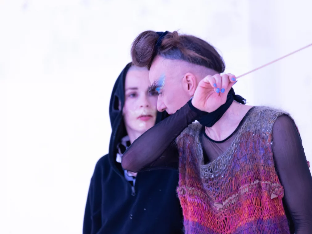 Zwei Performer*innen in einer hell ausgeleuchteten Szene. Eine Person mit hochgestecktem Haar, blauem Lidschatten und transparentem, buntem Oberteil hält einen dünnen Stab und hebt den Arm. Dahinter steht eine Person mit Kapuzenpullover.