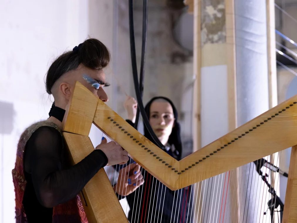 Eine Person mit auffälligem Make-up spielt eine große Harfe, während im Hintergrund eine weitere Person mit Kapuze und hellem Make-up zu sehen ist. Die Szene ist in kühles Licht getaucht, Kabel und Instrumente strukturieren den Raum.