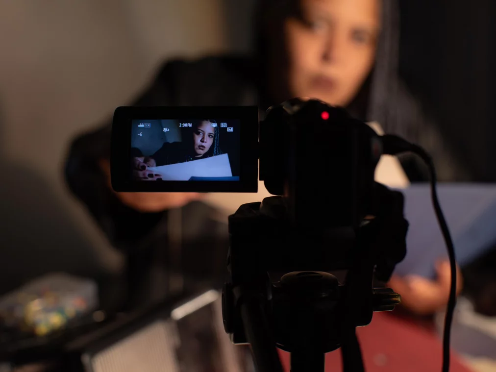 A person with dark lipstick and braided hair sits in front of a camera, holding papers in their hands. Their face is sharply visible on the camera’s display, while they appear blurred in the background. The scene is bathed in warm, subdued light.