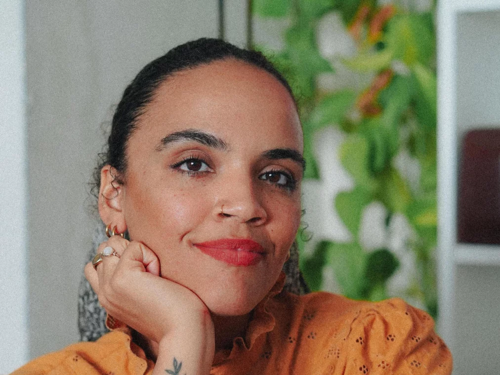 A person with red lipstick sits indoors, resting their head on their hand. They wear an orange embroidered blouse and have tattoos on their forearm. Green plants and bookshelves are visible in the background.