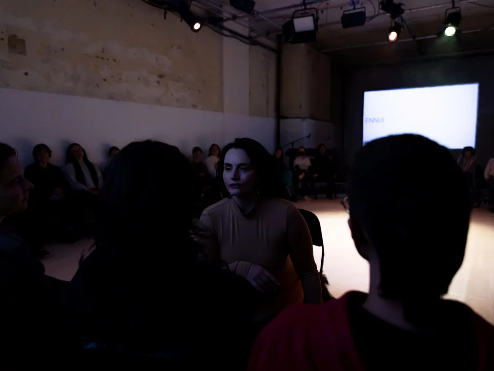Eine Person im hautfarbenen Ganzkörperanzug sitzt nah beim Publikum in einem abgedunkelten Raum. Im Hintergrund sind Zuschauende im Halbkreis und eine Leinwand mit dem Wort „ENNUI“ zu sehen
