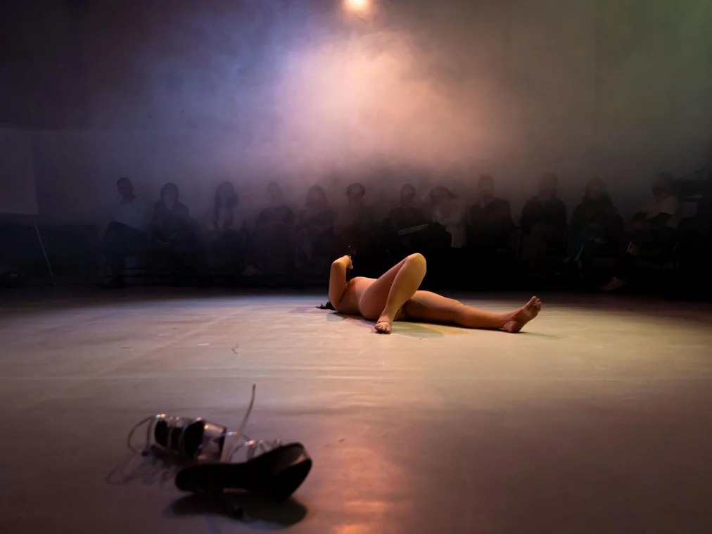 Eine Person liegt auf dem Bühnenboden in einer Rauch- und Lichtstimmung. Vor ihr liegen durchsichtige Plateauschuhe, im Hintergrund sitzt das Publikum im Dämmerlicht.