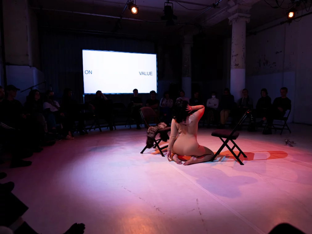Eine Person sitzt mit dem Rücken zum Publikum auf dem Boden zwischen zwei Stühlen. Hinter ihr zeigt eine Leinwand den Schriftzug „ON VALUE“.