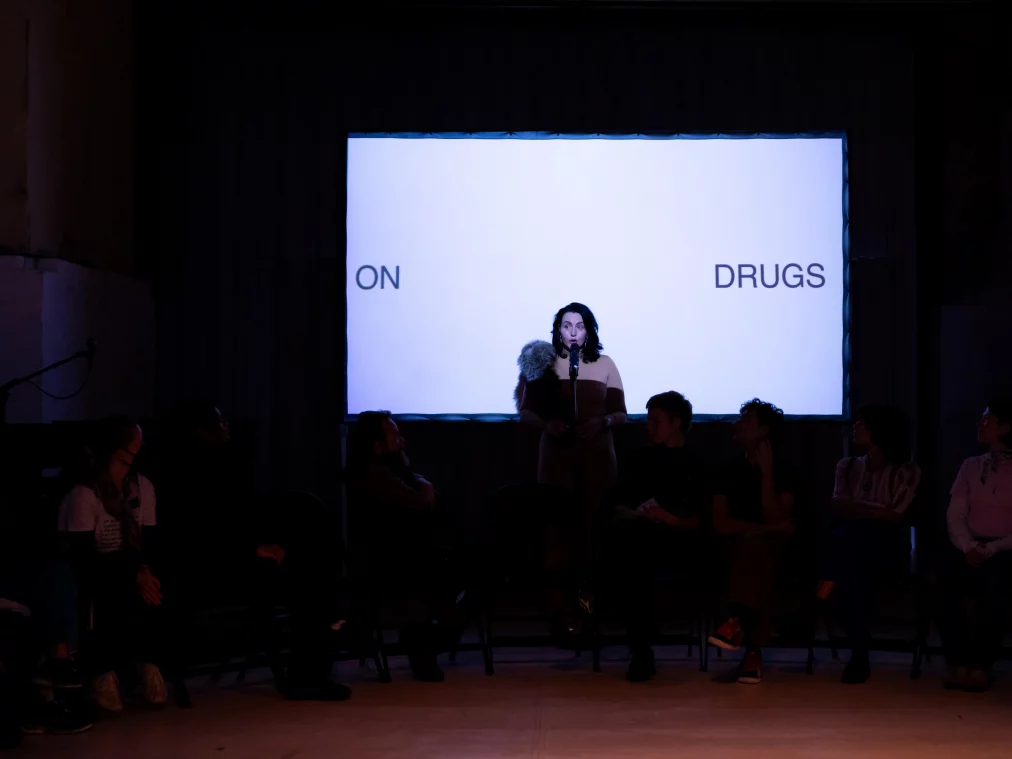 Eine Person steht auf einer Bühne vor einem großen Bildschirm mit der Schrift „ON DRUGS“. Sie spricht in ein Mikrofon, während Menschen im Hintergrund sitzen und zusehen.