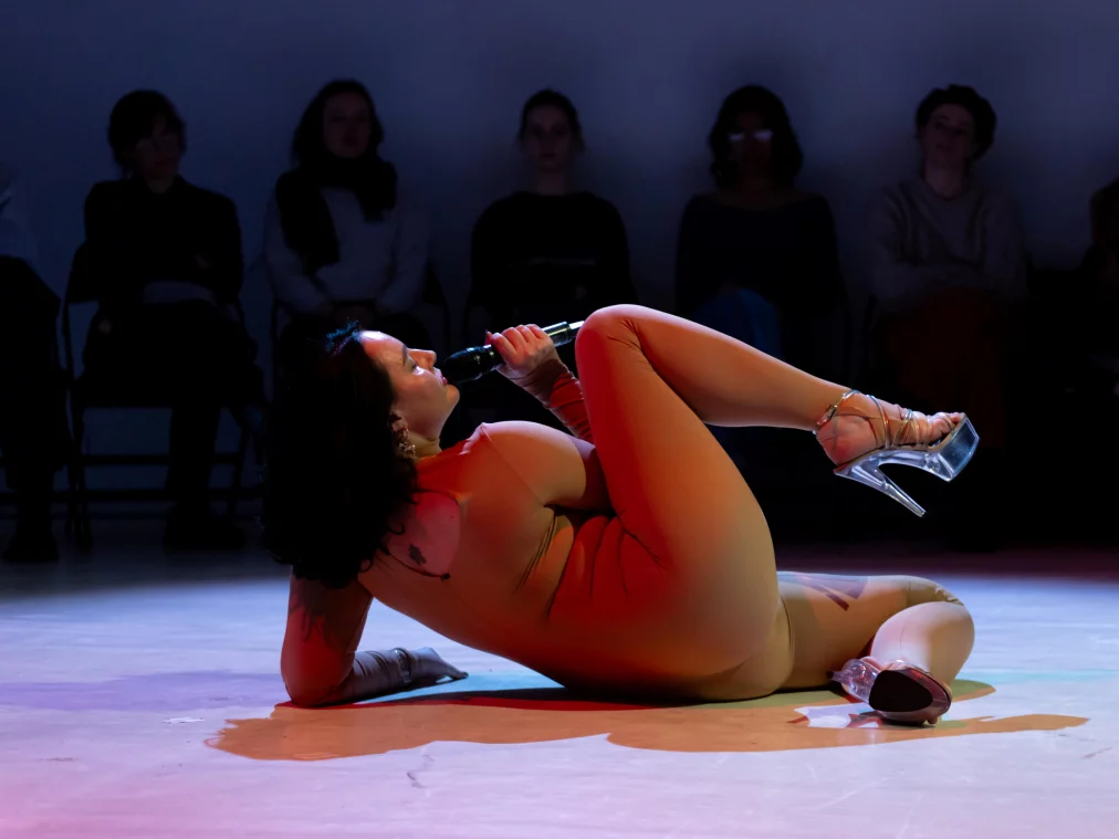 Eine Person im hautfarbenen Ganzkörperanzug liegt auf dem Boden, hält ein Mikrofon und blickt ins Publikum. Im Hintergrund sitzen Zuschauende im Halbkreis.