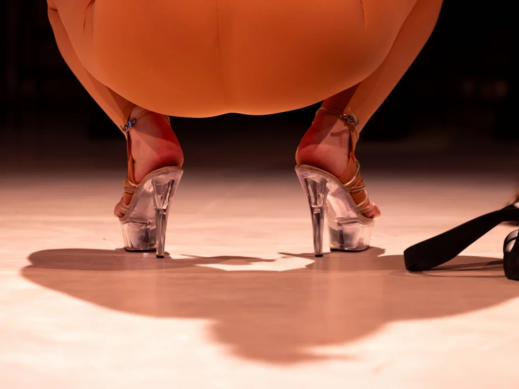 Nahaufnahme von Füßen in durchsichtigen Plateauschuhen mit hohen Absätzen. Die Person trägt einen hautfarbenen Ganzkörperanzug und hockt auf dem Boden, neben ihr liegt eine schwarze Tasche.