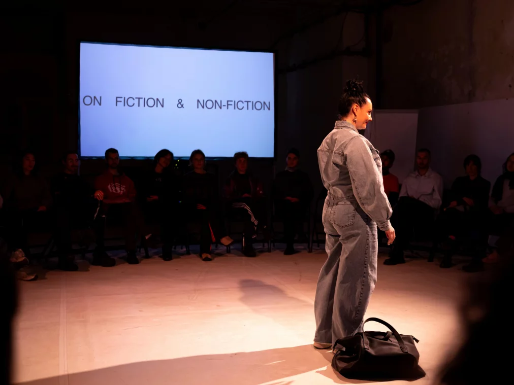 Eine Person in einem grauen Jeans-Overall steht im Lichtkreis auf der Bühne. Neben ihr liegt eine schwarze Tasche, im Hintergrund sitzt das Publikum. Auf einer Leinwand steht „ON FICTION & NON-FICTION“.