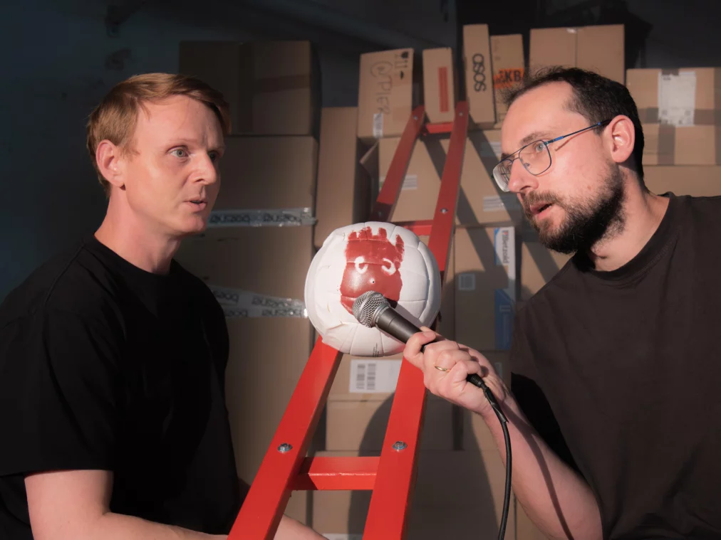 Two people sit in a room filled with cardboard boxes. Between them, a white volleyball with a painted face rests on a red ladder, with a microphone pointed toward it.