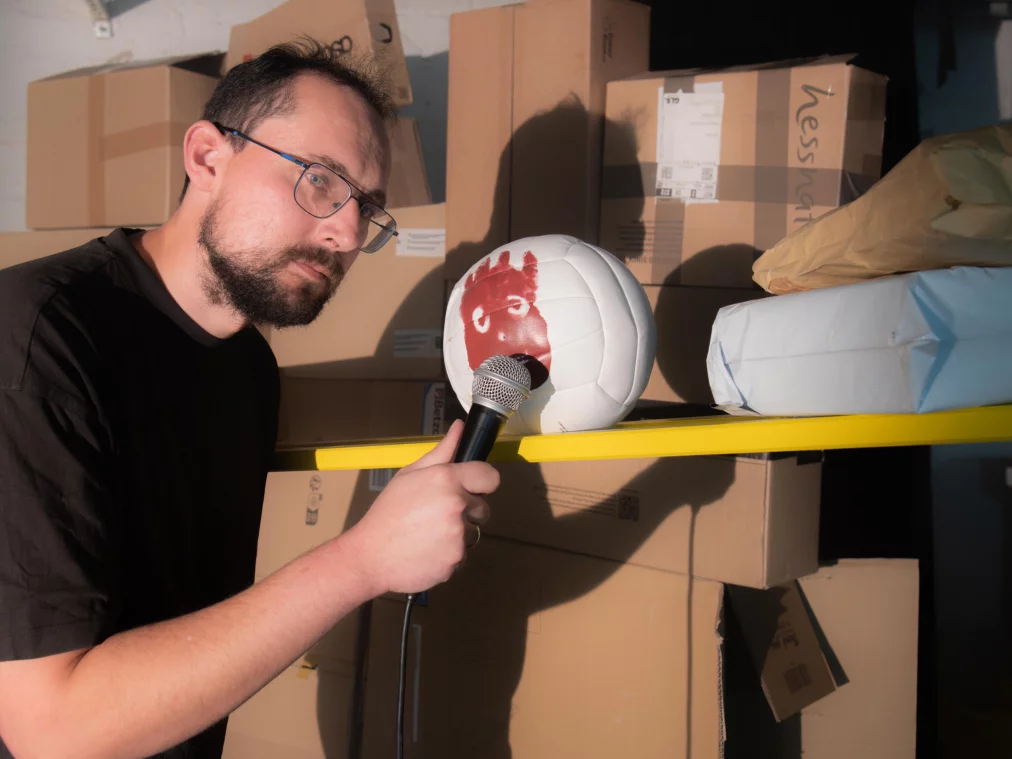 A person holds a microphone to a white volleyball with a painted face, resting on a yellow shelf. Behind them, several cardboard boxes are stacked.