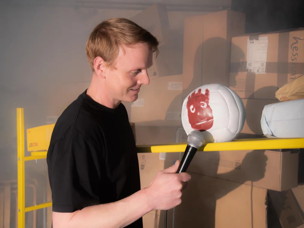 A person holds a microphone in front of a white volleyball with a painted face, placed on a yellow shelf. Stacked cardboard boxes are visible in the background.