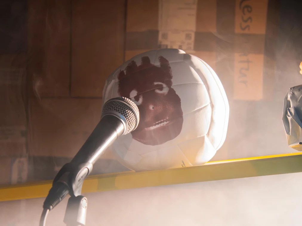A microphone is pointed at a white volleyball with a painted face, lying on a yellow bar. Cardboard boxes and mist are visible in the background.