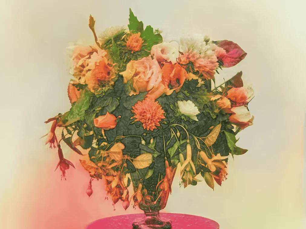 Ein Blumenarrangement in warmen Orange- und Rottönen steht auf einem pinken Podest. Die Blumen wirken leicht welk und sind von einem weichen, nebligen Licht umgeben.