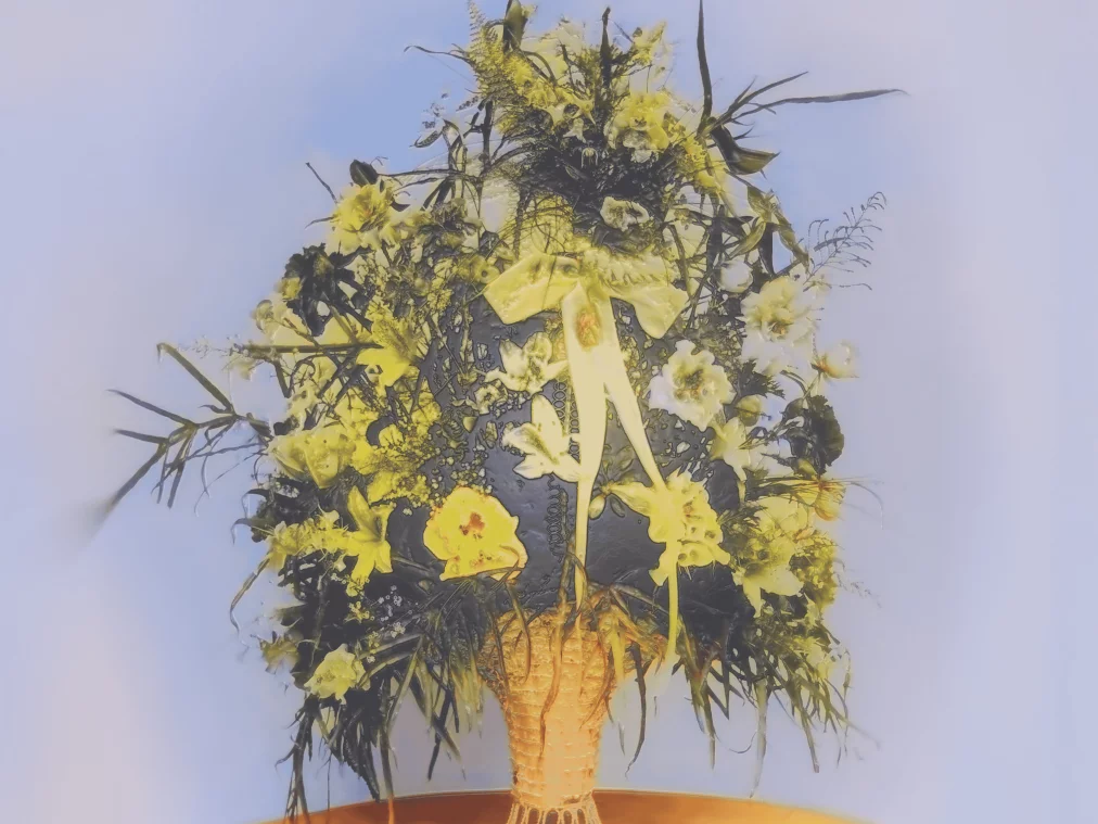 Ein großes, gelb-weißes Blumenarrangement mit Bändern steht in einem geflochtenen Korb auf einem gelben Tisch, vor einem hellblauen Hintergrund.