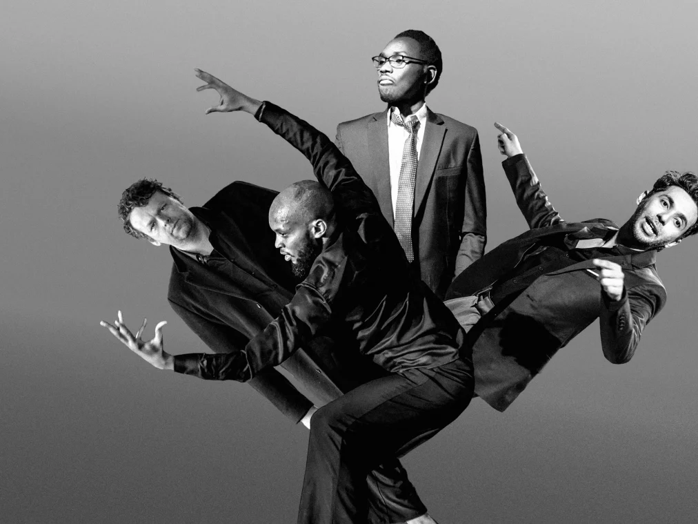 Four people stand and lean in a dynamic, collage-like arrangement. All are dressed in suits. The person in front bends forward with an expressive arm gesture. To the left, another person tilts slightly backward. Behind them, one person stands upright and calm, while the person on the far right leans back with an outstretched pointing arm. The black-and-white image conveys a theatrical and energetic atmosphere.