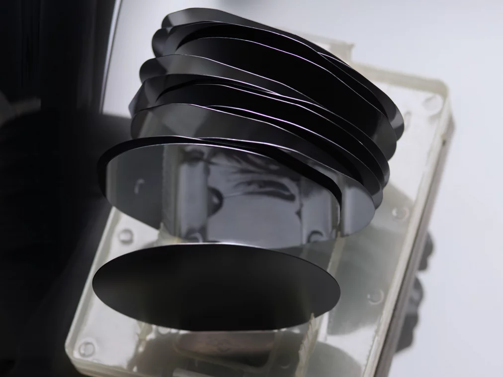 Several stacked black silicon wafers in a transparent holder; their reflective surfaces cast soft reflections and distort shapes in the background.