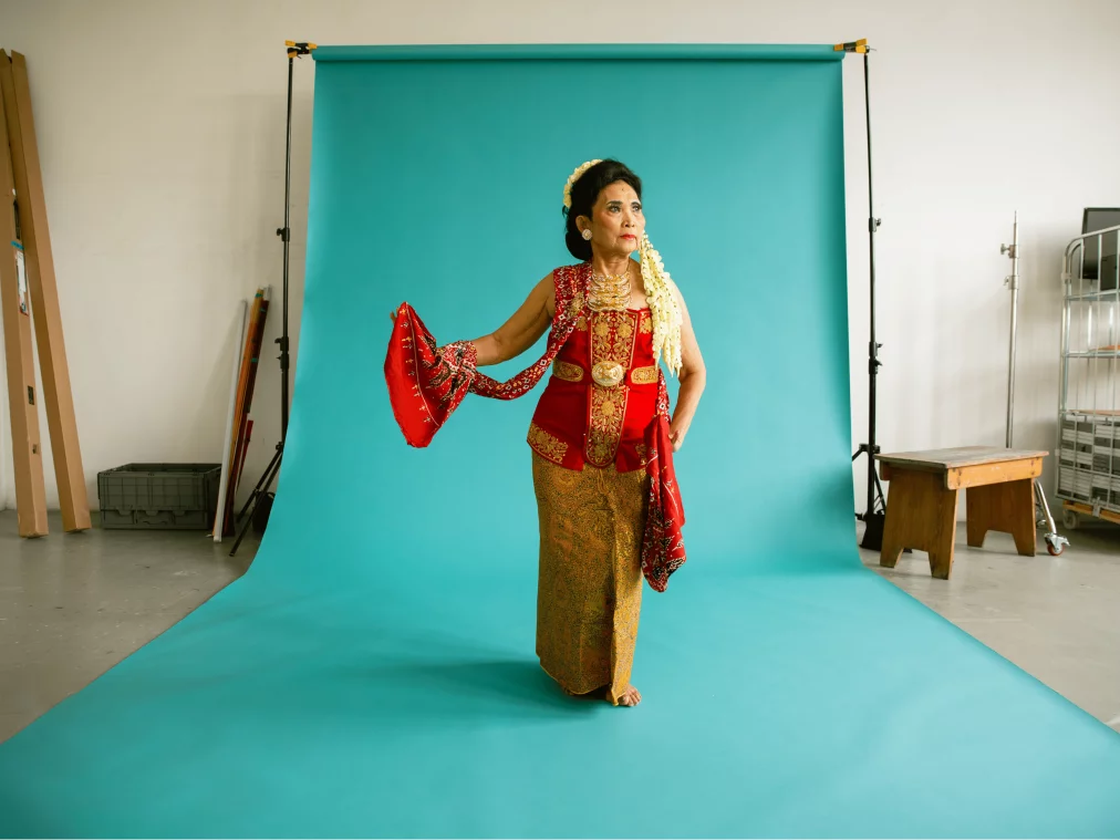 Eine ältere Tänzerin steht barfuß vor einem türkisfarbenen Studiohintergrund und trägt ein traditionelles indonesisches Gewand in Rot und Gold. Sie hält ein rotes Tuch elegant zur Seite und blickt konzentriert nach vorne.
