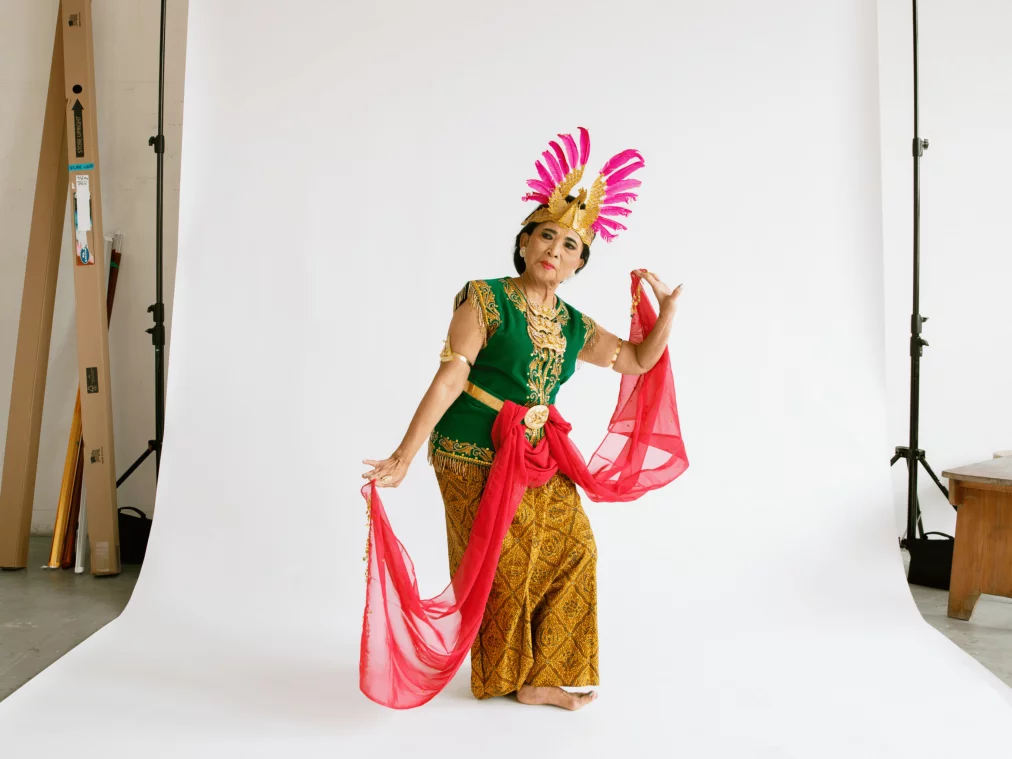 Eine ältere Tänzerin in farbenfrohem traditionellen Kostüm mit grünem Oberteil, goldenen Mustern und pinkem Kopfschmuck posiert vor einem weißen Studiohintergrund. Sie hält ein langes pinkes Tuch in beiden Händen und steht barfuß.