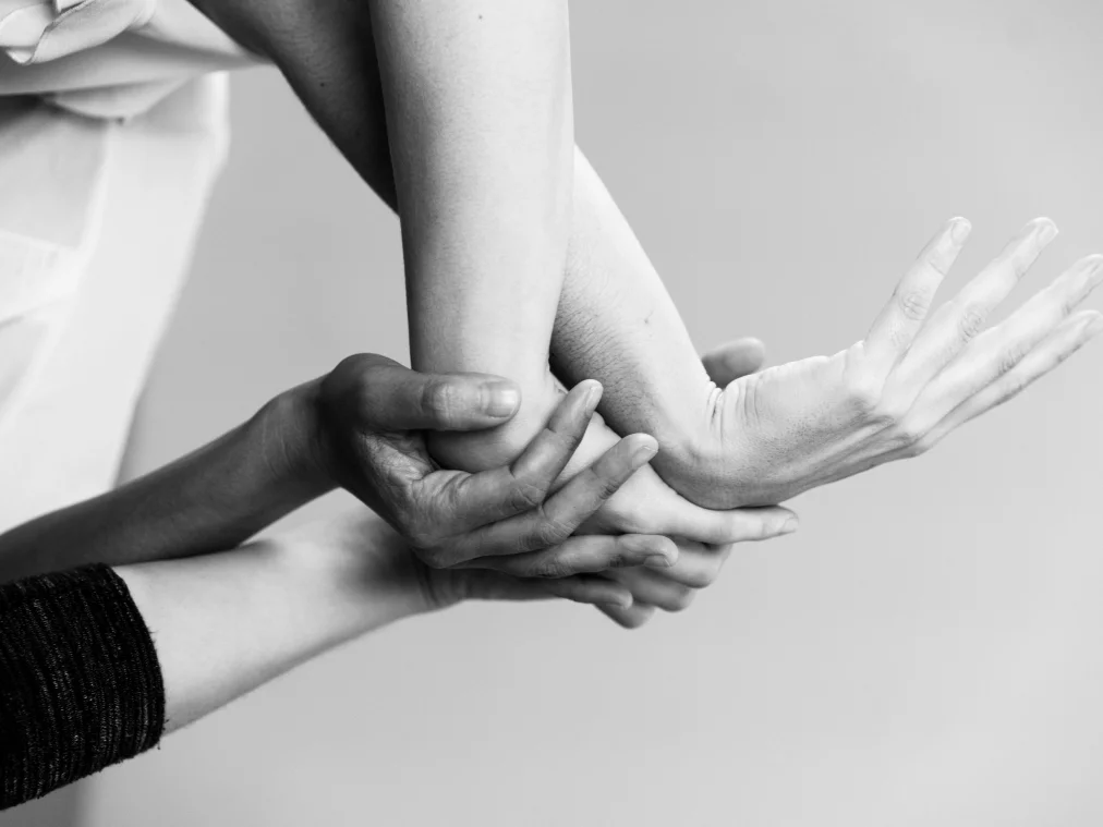 Nahaufnahme mehrerer Hände, die einen ausgestreckten Unterarm halten und unterstützen. Die Finger greifen vorsichtig ineinander; die Szene wirkt konzentriert und sanft zugleich.