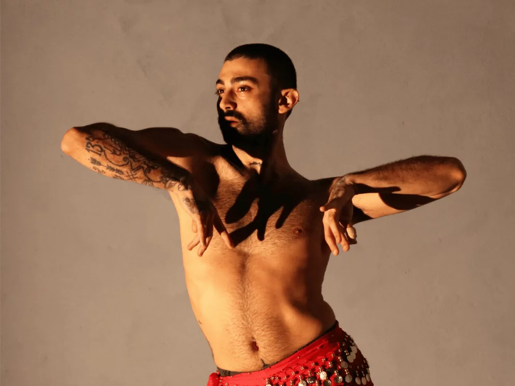 Eine tanzende Person mit nacktem Oberkörper und roten, bestickten Hüfttuch posiert in elegant geschwungenen Armformen. Tätowierungen bedecken die Arme, und warmes Licht erzeugt starke Schatten auf Haut und Hintergrund.