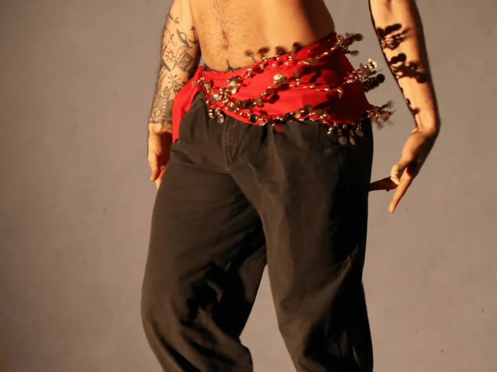 Nahaufnahme eines tanzenden Oberkörpers, bekleidet mit einer schwarzen Hose und einem roten Hüfttuch mit glänzenden Münzen und Perlen, die beim Bewegen schimmern. Tätowierte Arme rahmen die Szene, während warmes Licht Schatten auf den Körper wirft.