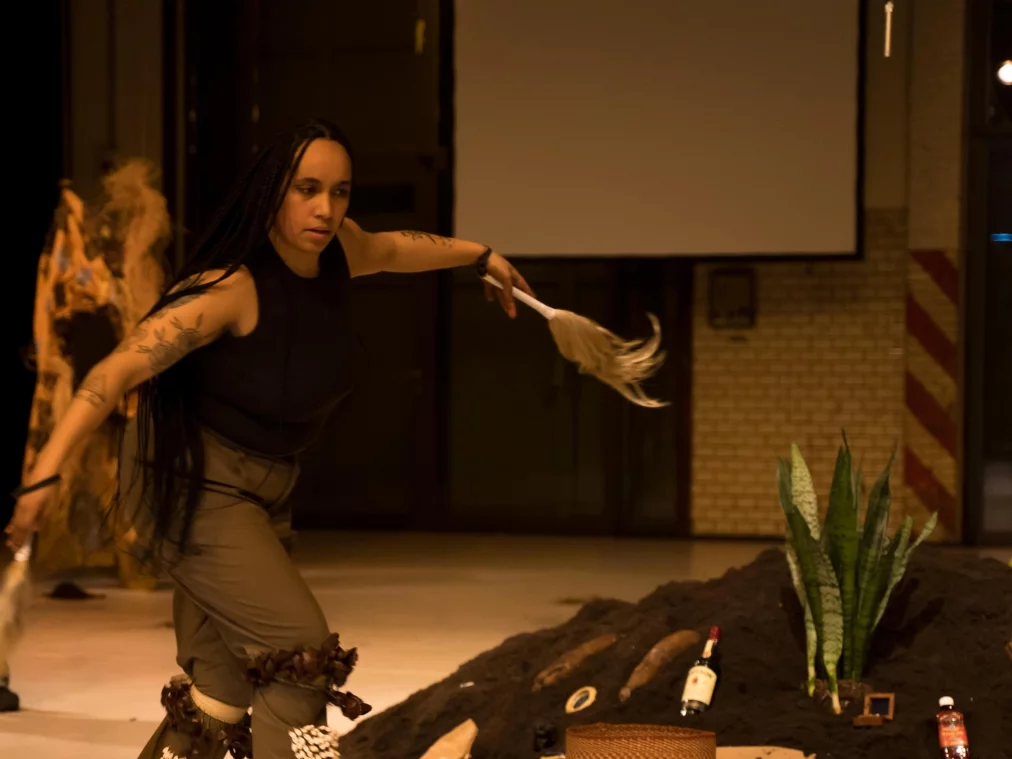 Eine Performer*in mit langen, geflochtenen Haaren bewegt sich tanzend über die Bühne und hält ein weißes Federbusch-Objekt in der ausgestreckten Hand. Neben ihr liegt ein großer Erdhügel, darauf arrangierte Objekte wie Flaschen, Pflanzen, Holzstücke und ein Schild mit der Aufschrift „ANCESTORS“. Die Szene ist warm ausgeleuchtet.