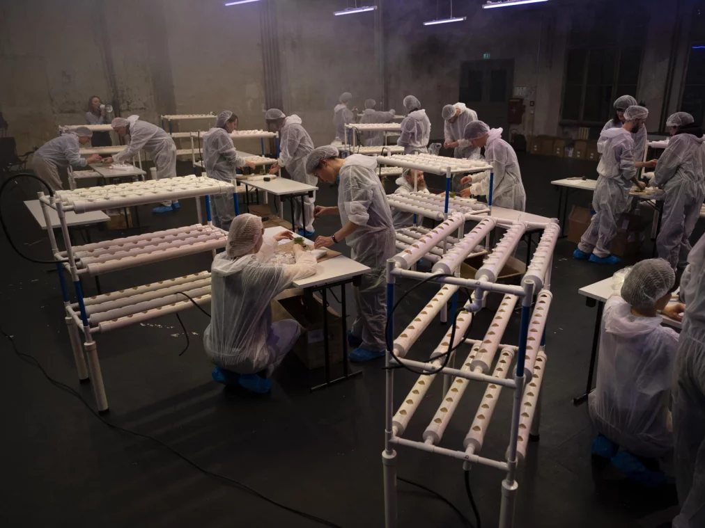 Mehrere Personen in weißen Schutzanzügen, Haarnetzen und blauen Überschuhen arbeiten in einem großen, leicht vernebelten Raum an langen Tischen und modularen PVC-Gestellen. Die Szene wirkt wie ein steriler Produktions- oder Laborprozess in einem performativen Setting.