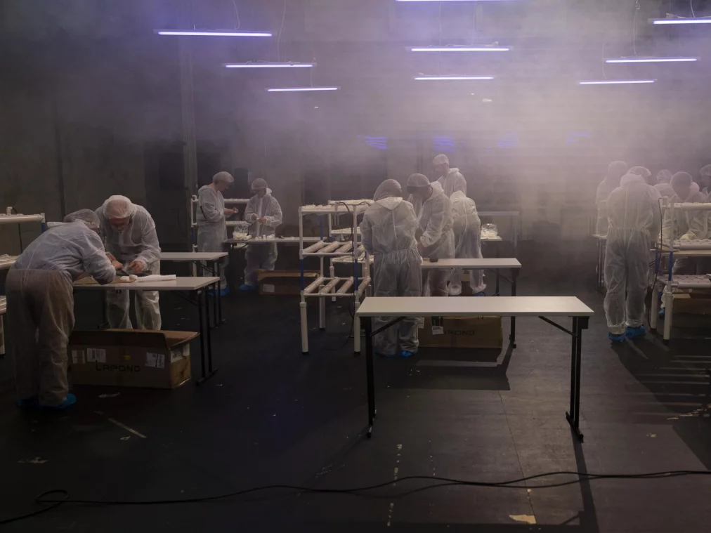 Eine größere Gruppe von Menschen in weißen Schutzanzügen verteilt sich in einem nebligen, industriellen Raum und arbeitet konzentriert an Tischen und PVC-Gestellen. Über ihnen hängen mehrere lineare LED-Leuchten, die ein kaltes, technisches Licht erzeugen.