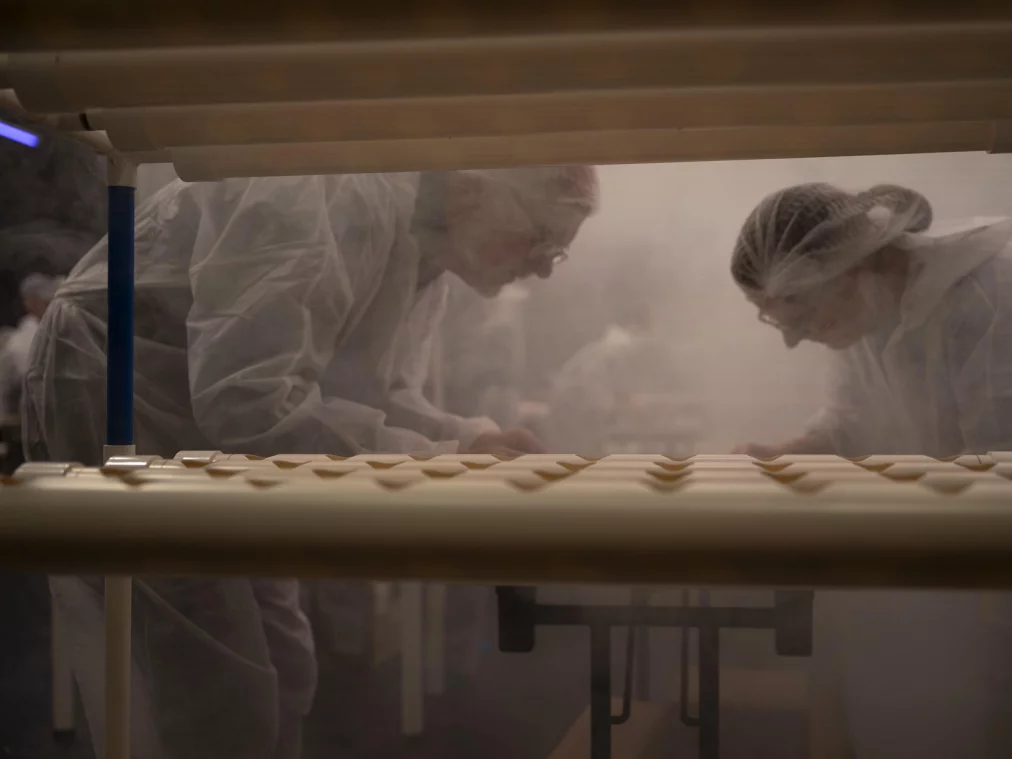 Durch die untere Ebene eines PVC-Rohrgestells hindurch blickt die Kamera auf zwei Personen in weißen Schutzanzügen, die sich über ihre Arbeit beugen und konzentriert an einem Bauteil arbeiten. Leichter Nebel erfüllt den Raum.