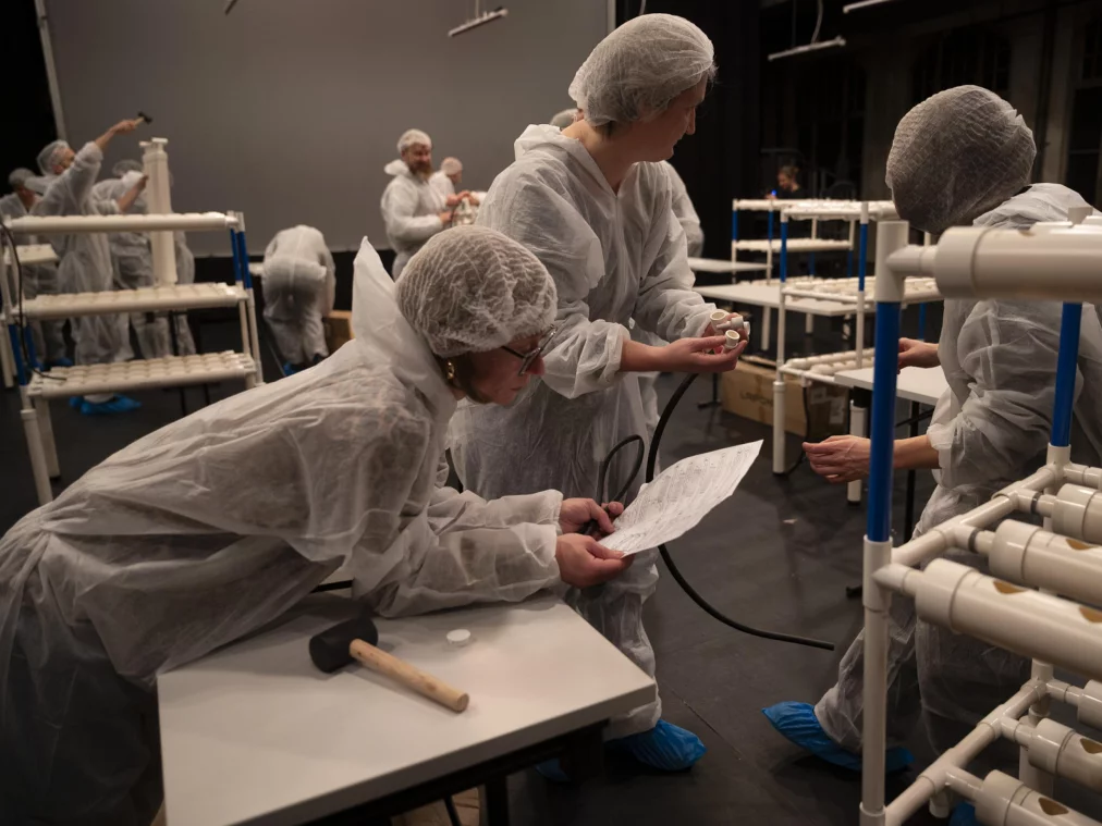 Eine Gruppe von Personen in weißen Schutzanzügen und Haarnetzen baut an Tischen ein Rohrsystem aus PVC-Teilen zusammen. Im Vordergrund lehnt sich eine Person über einen Tisch und hält eine Anleitung, während zwei weitere Personen Bauteile vergleichen.