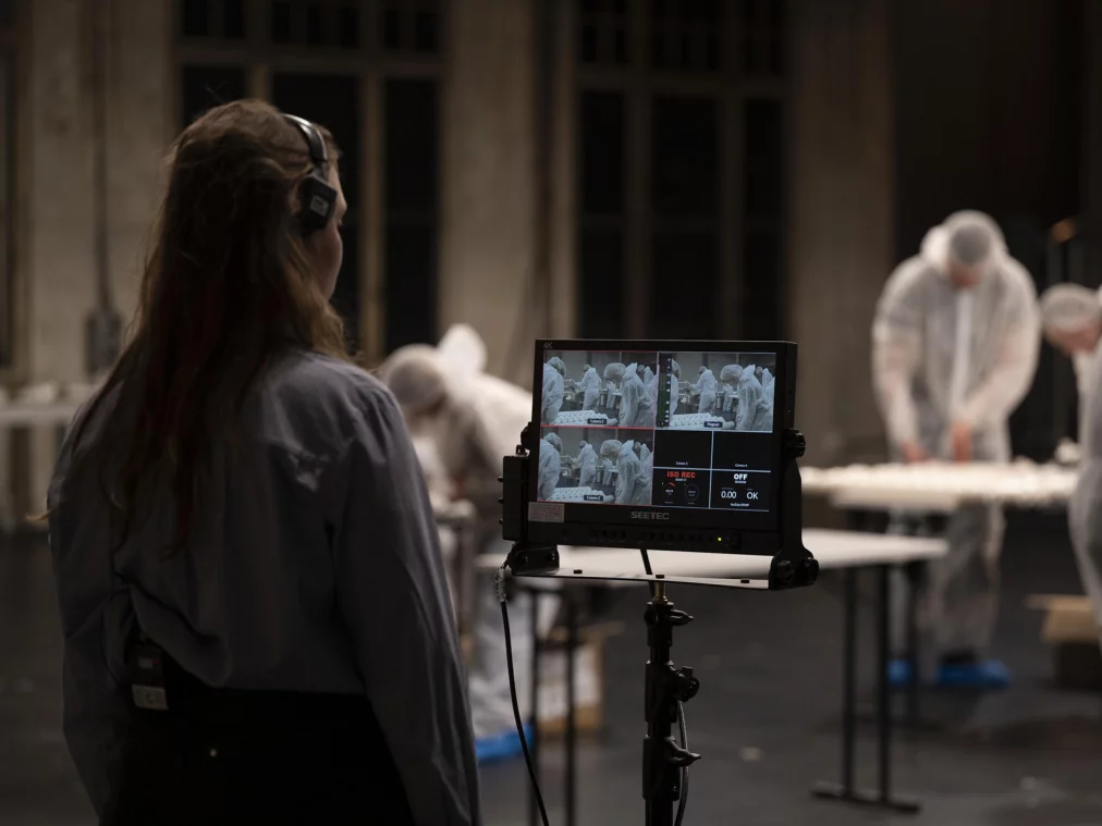 Eine Person mit langen Haaren steht mit Headset vor einem Monitor auf einem Stativ. Auf dem Bildschirm sind mehrere Kameravorschauen zu sehen, die Personen in Schutzanzügen bei einer Montagearbeit zeigen.