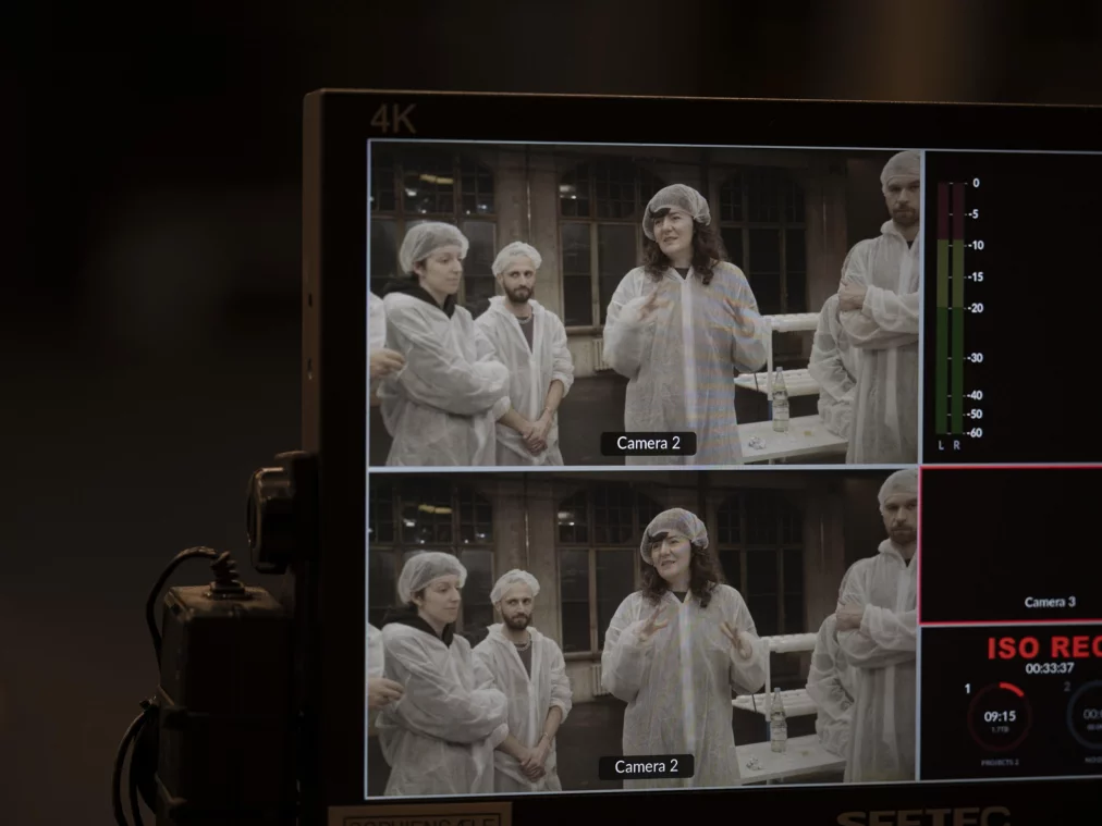 Ein Monitor zeigt zwei Kameraansichten derselben Szene: mehrere Personen in Schutzanzügen stehen zusammen und eine Person in der Mitte spricht gestikulierend.