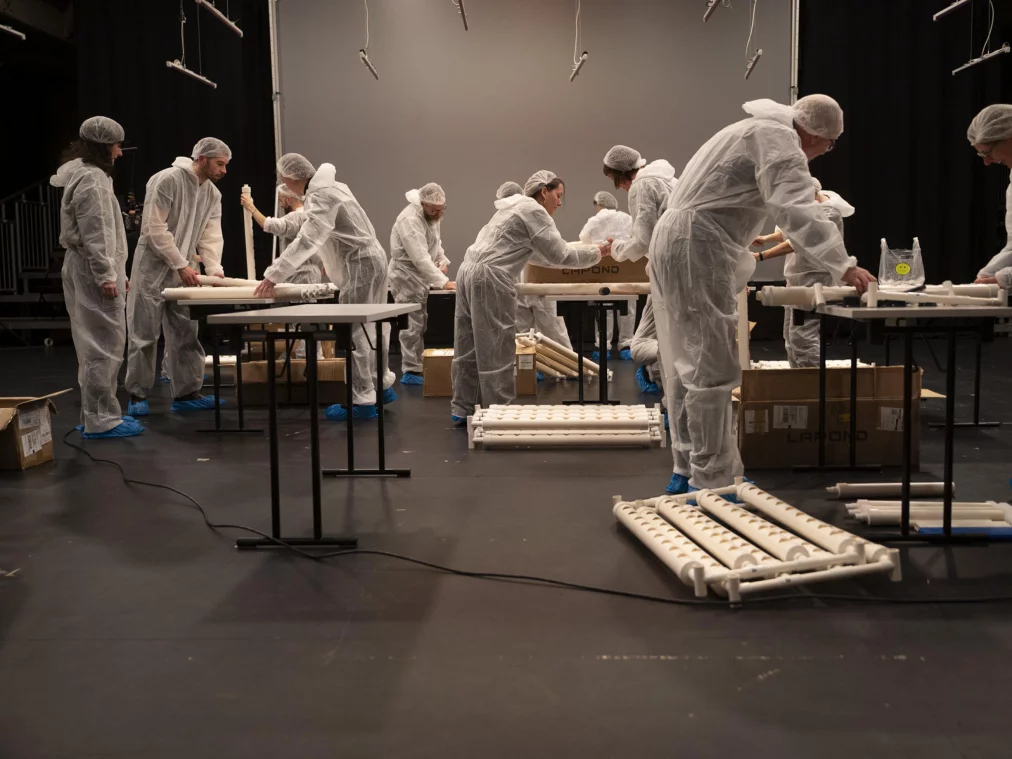Personen in weißen Schutzanzügen bauen an Tischen einzelne Module eines PVC-Gestells zusammen. Auf dem Boden stehen mehrere geöffnete Kartons mit Bauteilen.