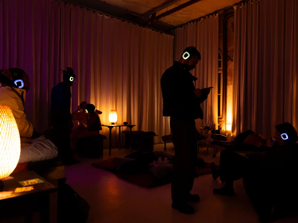 In einem abgedunkelten Raum sitzen und stehen mehrere Personen* mit leuchtenden Kopfhörern. Warmes, orange-violettes Licht fällt auf Sitzkissen, kleine Tische und weiche Stoffe. Die Atmosphäre wirkt ruhig und konzentriert.