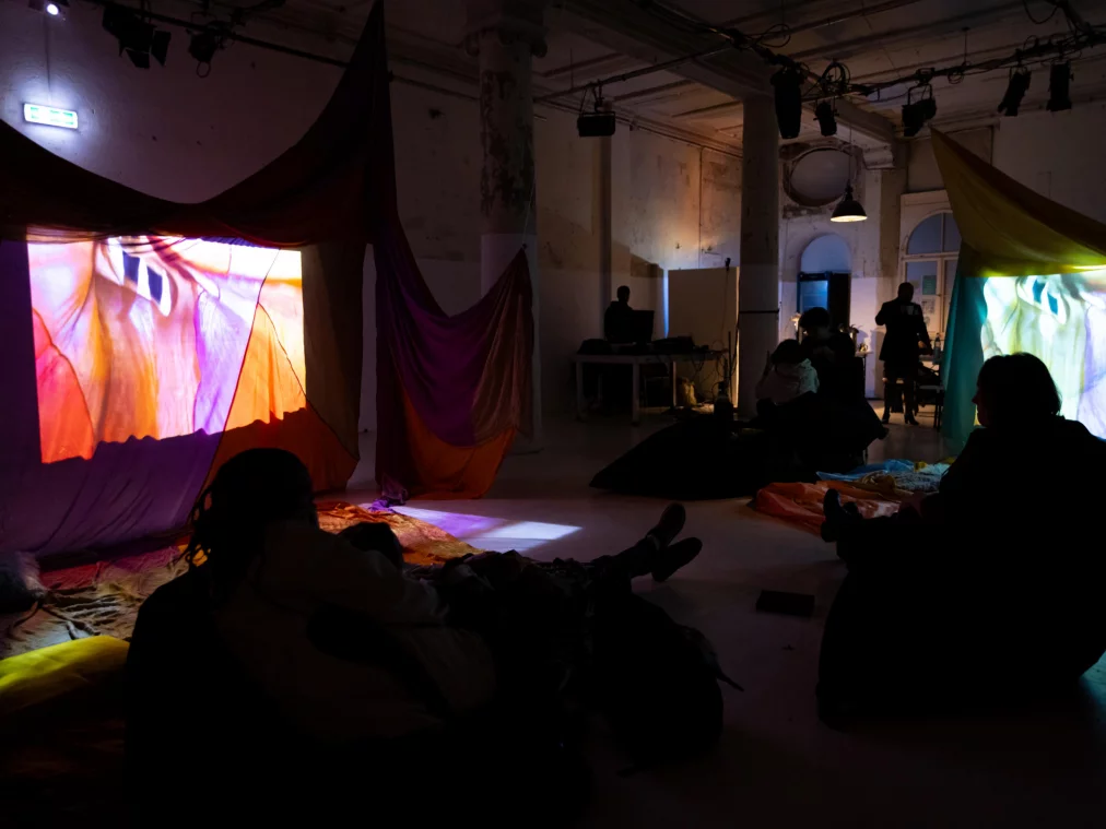 Menschen sitzen oder liegen entspannt in einem großen Raum, in dem bunte Stoffbahnen aufgehängt sind. Zwei projizierte, abstrakte Video-Bilder leuchten auf den Stoffen. Die Umgebung ist dunkel, nur von den Projektionen erhellt.