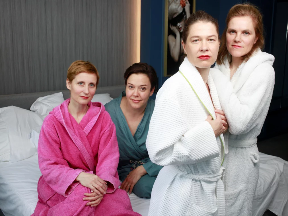 Vier Erwachsene sitzen und stehen in einem hellen Schlafzimmer, alle in farbigen Bademänteln. Zwei Personen sitzen auf einem ungemachten Bett, eine in Pink und eine in Blau. Zwei weitere Personen in weißen Bademänteln stehen nah beieinander im Vordergrund. Alle blicken direkt in die Kamera, in einer ruhigen, konzentrierten Atmosphäre.