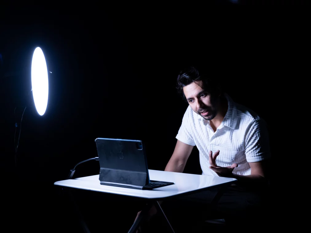 Eine Person sitzt an einem kleinen weißen Tisch in einem dunklen Raum. Ein starkes ringförmiges Licht beleuchtet das Gesicht, während die Person vor einem aufgeklappten Tablet spricht. Die Szene erinnert an eine Online-Kommunikationssituation.