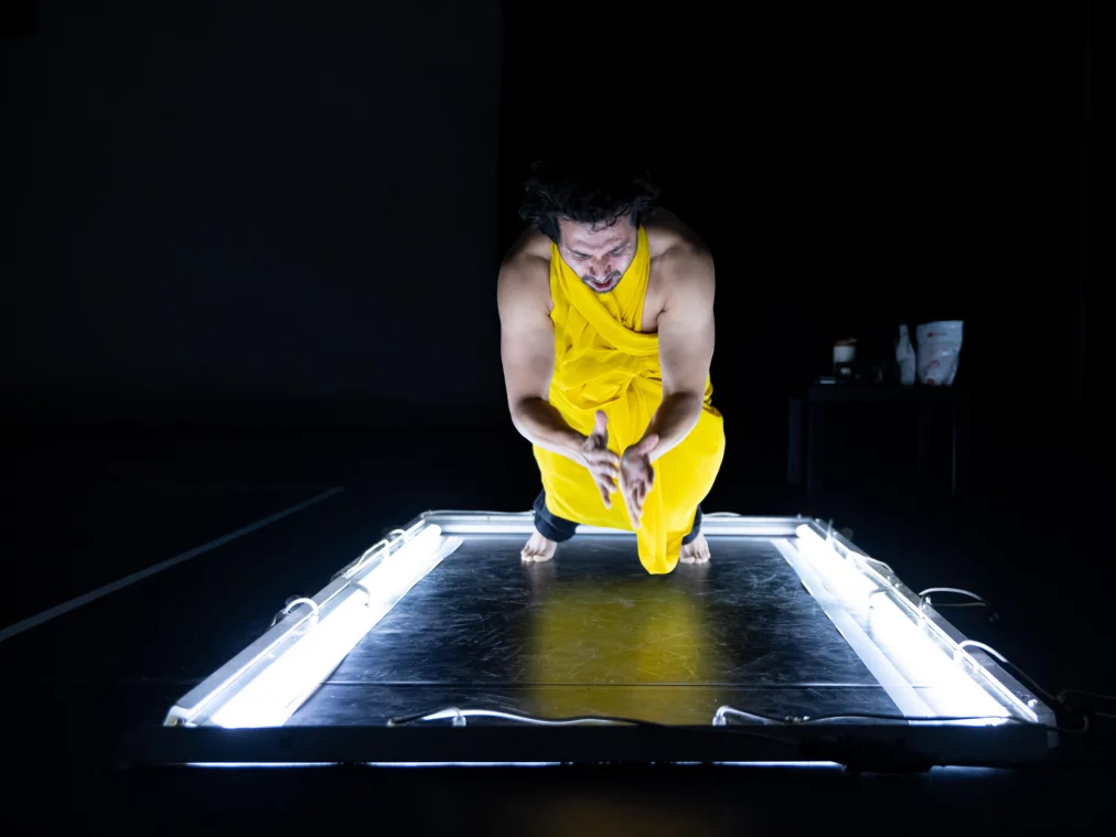 Eine Person in einem leuchtend gelben, drapierten Kostüm kniet auf einer von Neonröhren umrahmten Plattform. Die Person beugt sich nach vorn und führt die Hände klatschend zusammen, während der Hintergrund vollständig schwarz bleibt.